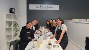 Five people in aprons smiling at a candle-making workshop inside a modern, grey-walled room labeled "banter & bliss candle co.