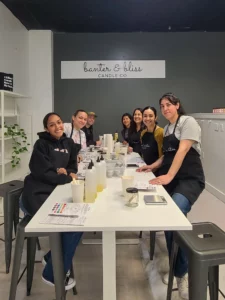 Group of people smiling at a candle-making workshop, sitting at a long table with craft supplies, in a room labeled "banter & bliss candle co.