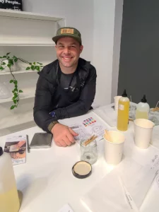 Man smiling at a table with samples and information papers, wearing a cap and black jacket, inside a bright room.