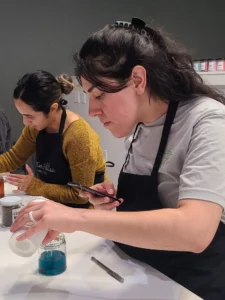 Two women in aprons making crafts, one using a smartphone, surrounded by jars of colorful pigments on a table.