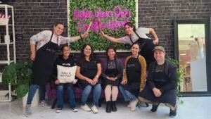 Group of seven people in aprons, toasting with drinks, smiling in front of a floral wall with neon sign "make your own coffee here".