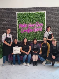 Group of seven smiling people in aprons posing in front of a wall with a green plant backdrop and a sign reading "make your own candle here!.