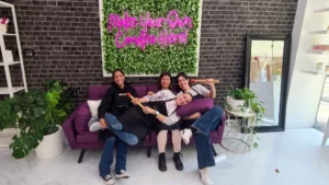 Four people smiling and posing together on a purple couch in a lobby with a neon sign that reads "make your own candle here!.