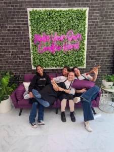 Four friends posing on a purple couch under a neon sign that reads "make your own candles here!" with a green leafy backdrop.